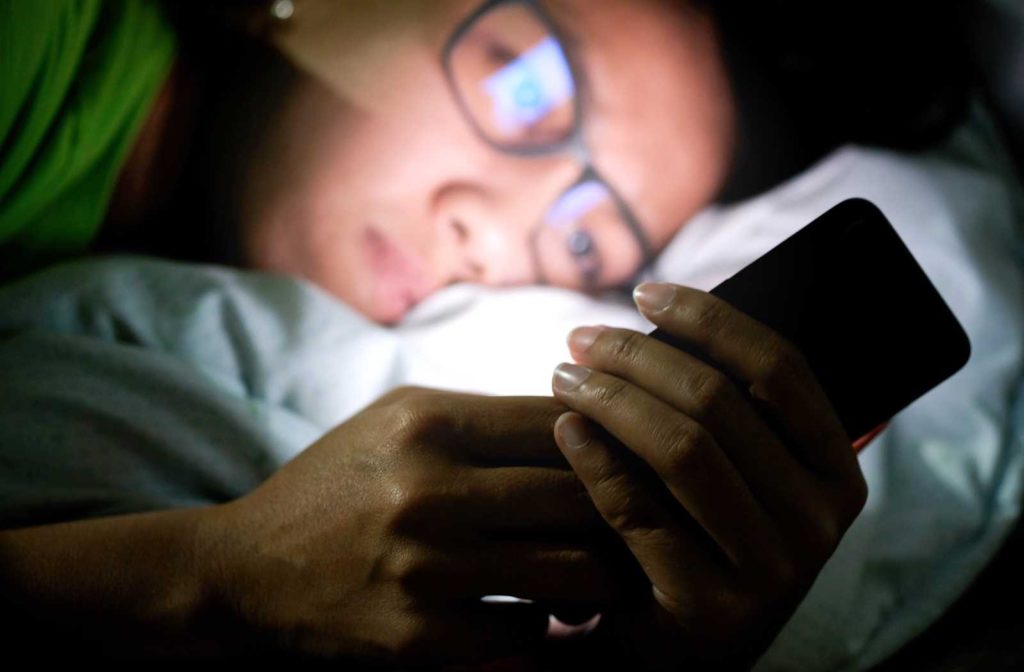 Woman on smartphone in dark bedroom with glasses reflecting blue light.