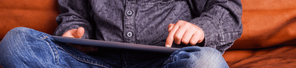 A cropped image of a young child wearing jeans and a dark denim button-up shirt, playing on a tablet in their lap, while sitting cross-legged on an orange couch