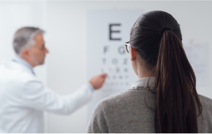 Female patient looking at eye chart on the wall while the male dentist points to the fourth row of letters on the chart.