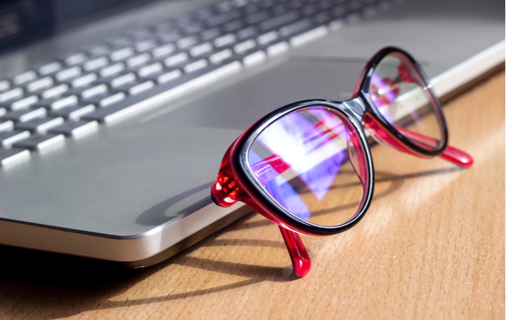 A pair of red glasses resting on the edge of a laptop with glare from sunlight through a nearby window visible on the lenses