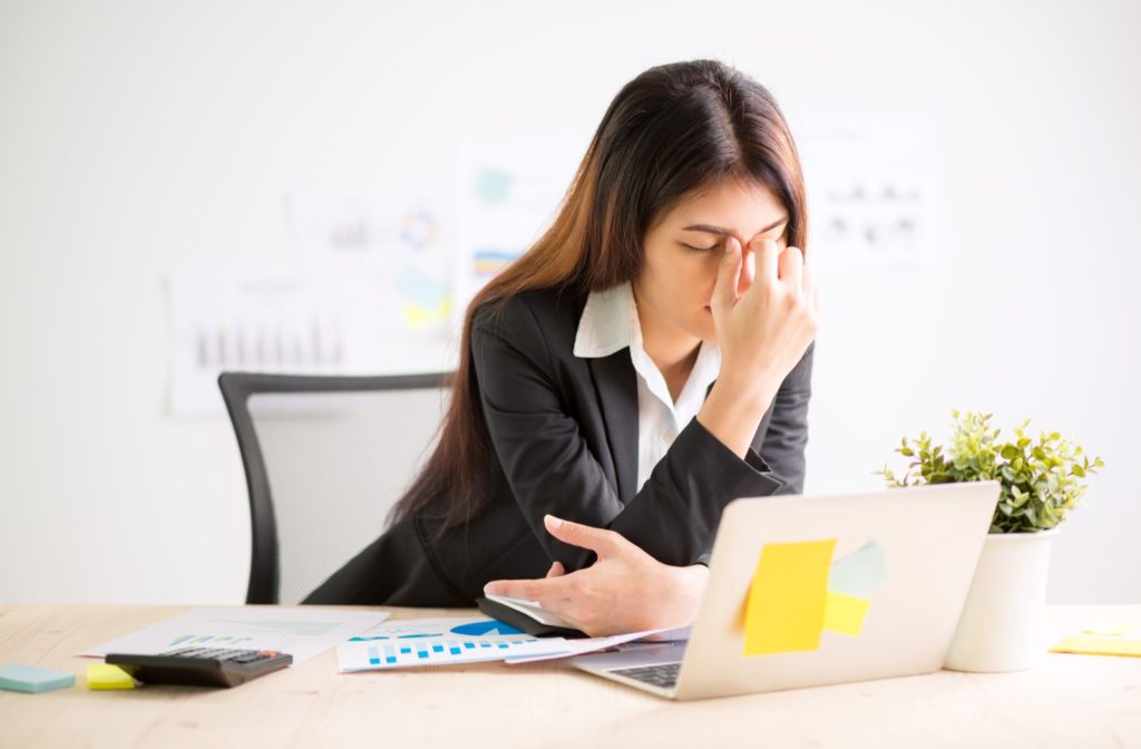 A businesswoman seated at her desk holding her eyes in discomfort from looking at her laptop screen for a prolonged period of time needing relief