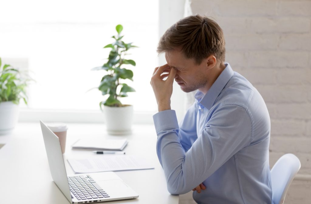 A side view of a man sitting at a desk with his laptop, holding both of his eye's in discomfort due to his digital eye strain