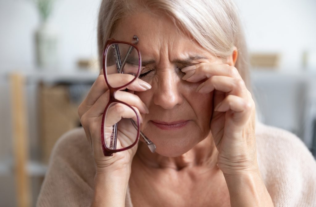 An older woman holding her glasses in her right hand, using both of her hands to rub her eyes due to the discomfort from dry eye