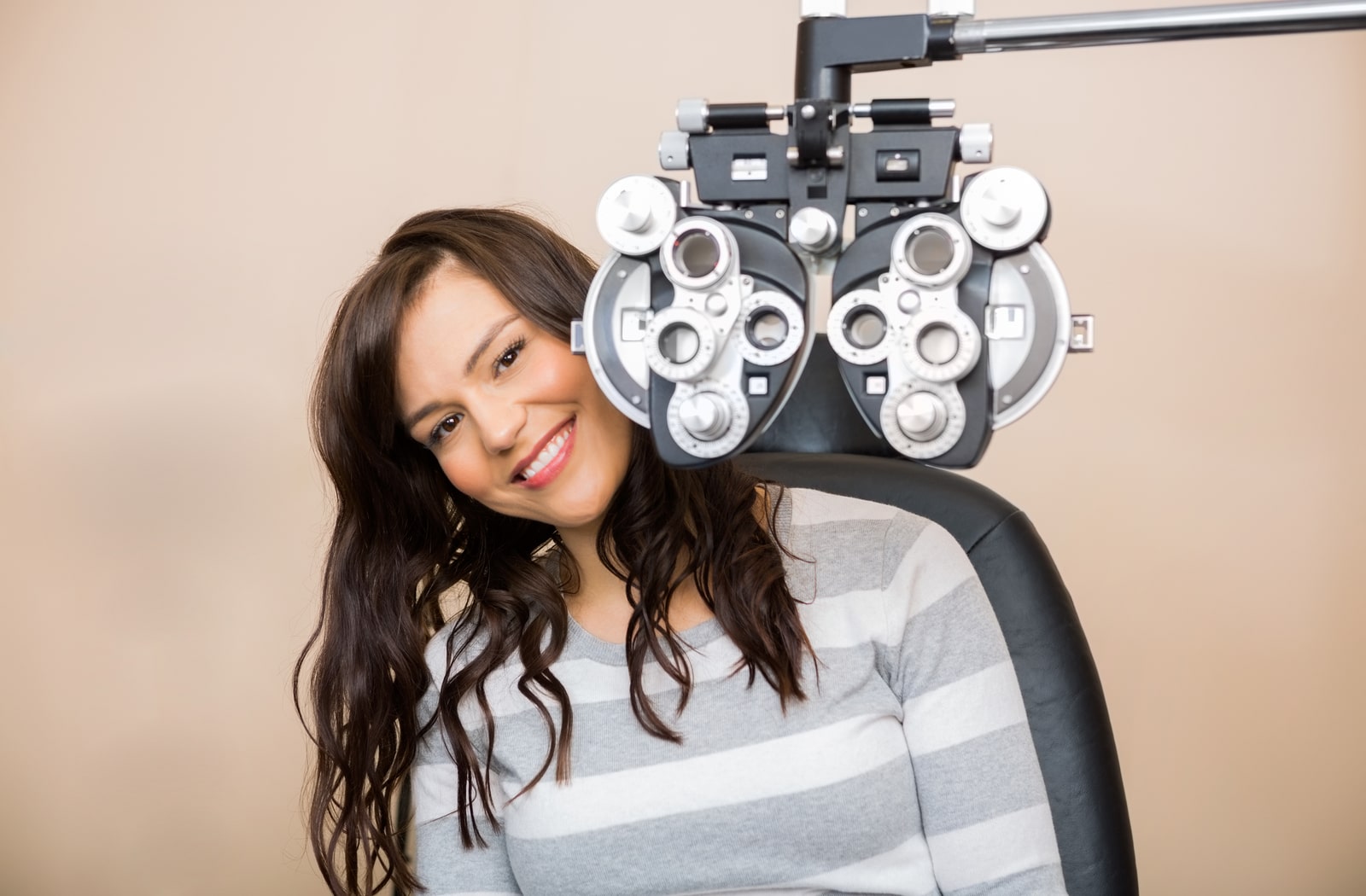 A woman sitting behind a phoropter at an optometrist office, smiling