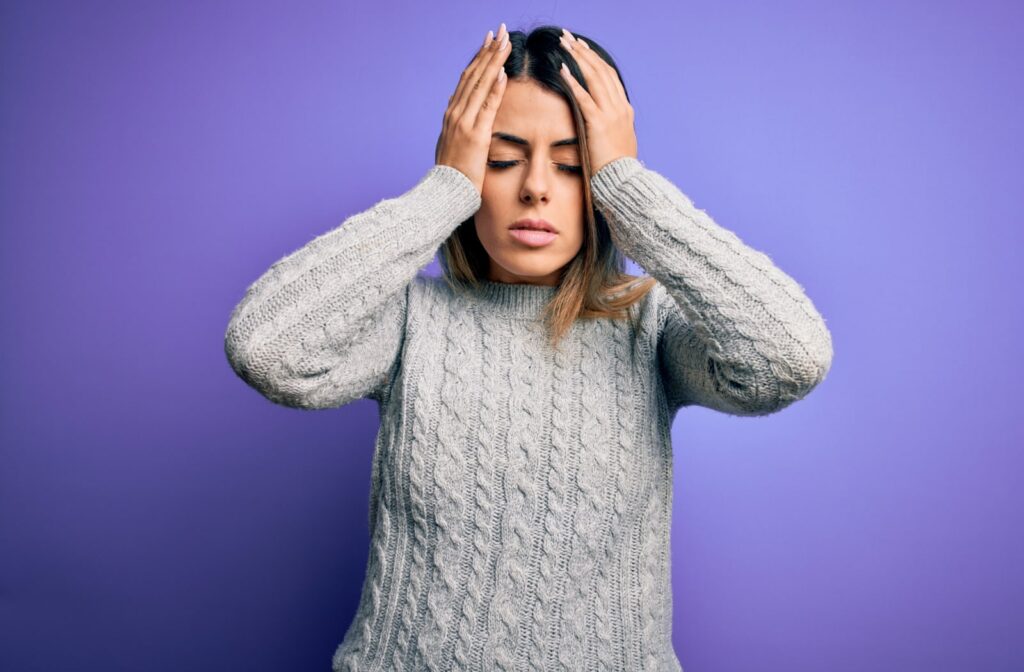 A woman standing in front of a purple background, and she's holding onto her head due to headaches.