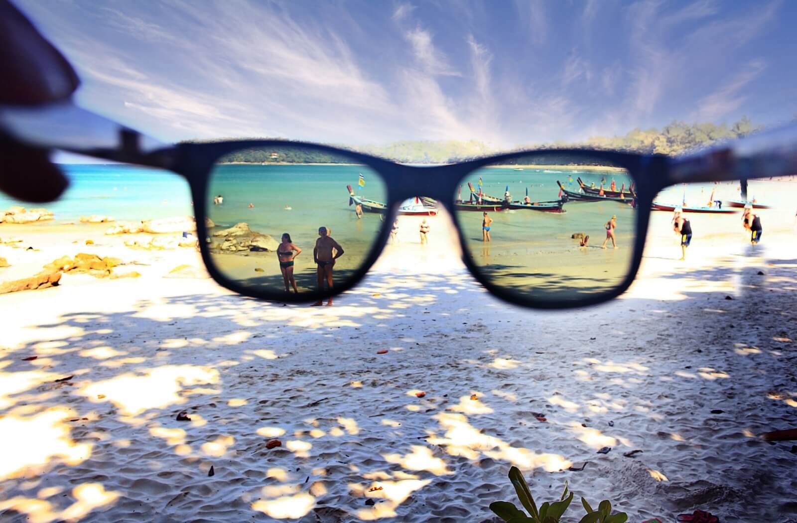 A hand holding up a pair of polarized sunglasses toward a beach full of people and boats.