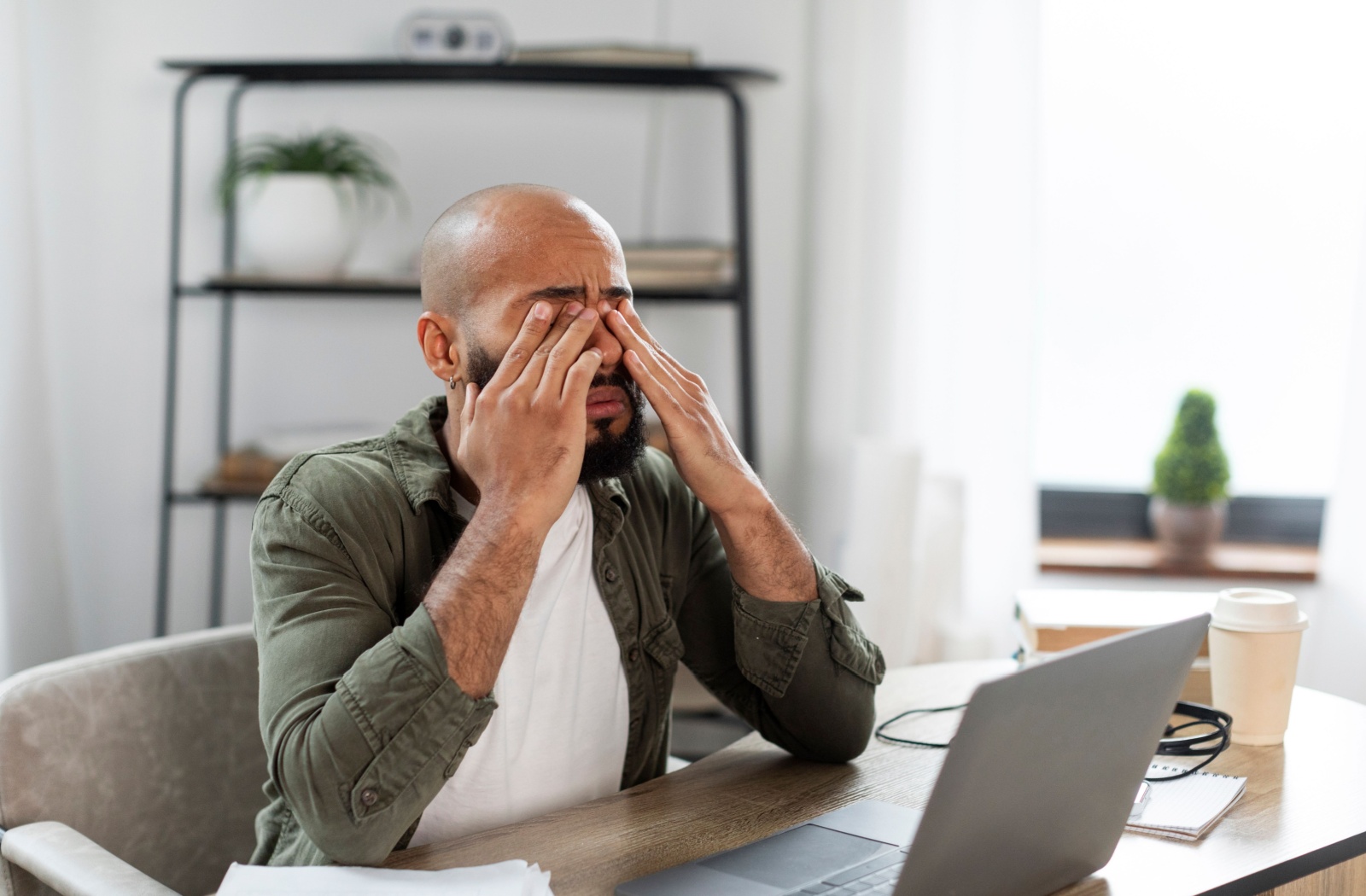 A person is sitting at a desk wearing a white t-shirt with a green shirt over top and they are rubbing their eyes with both hands and look uncomfortable.