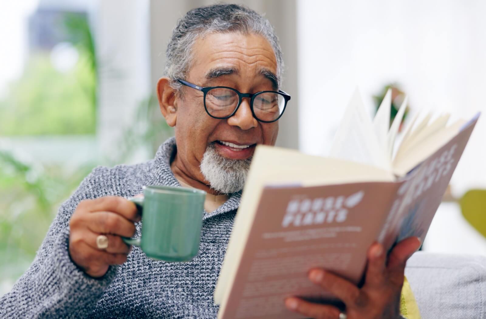 In a well-lit room, an older adult enjoys a book and a warm beverage, using reading glasses to comfortably manage age-related presbyopia.